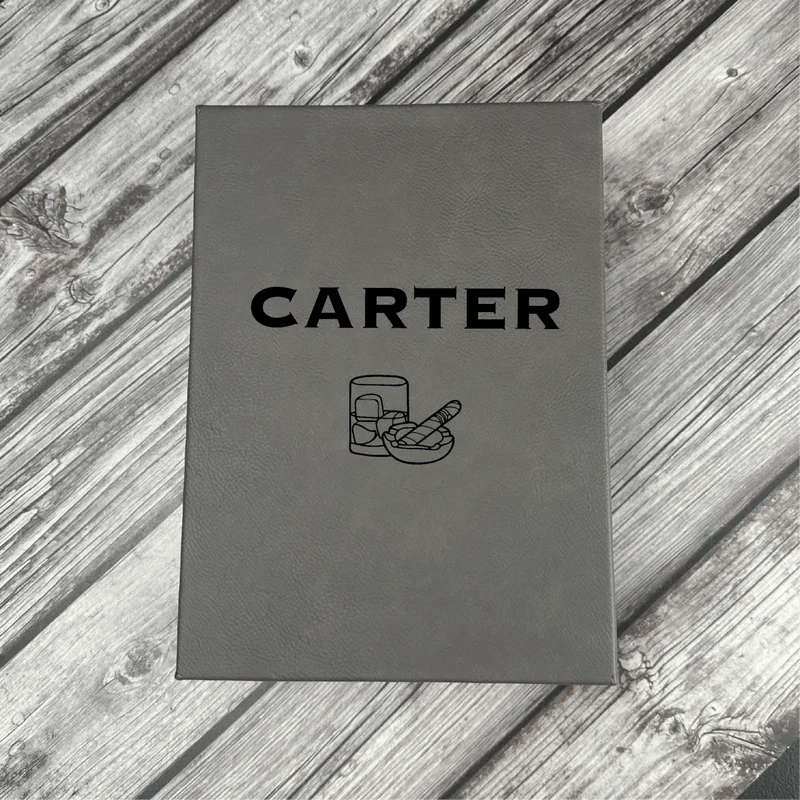 Gray leatherette box with "CARTER" engraved on cover and flask and cigar icons below, displayed on weathered wood surface