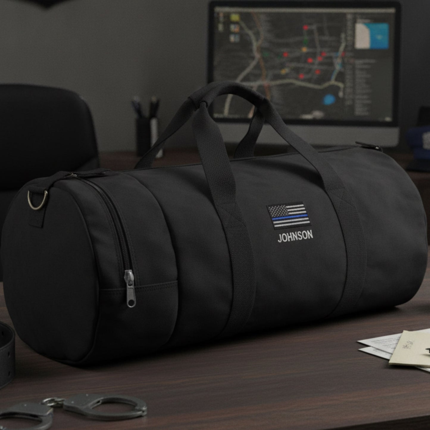 Black cylindrical duffle bag with sample personalization showing "JOHNSON" text and thin blue line flag emblem on front.