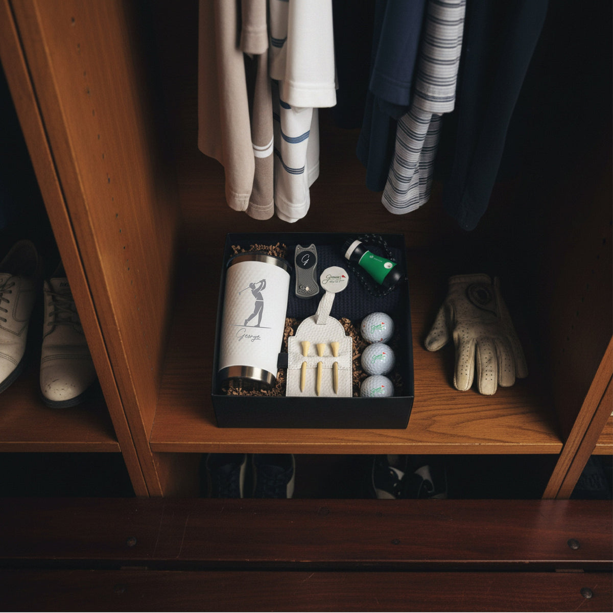 Golf-themed gift box with engraved flask, divot tool, golf balls, and accessories displayed on wooden shelf with clothing and shoes.