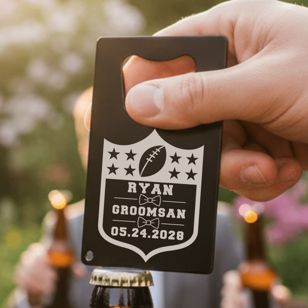 Hand holding a personalized bottle opener with 'Ryan Groomsman' text against a blurred outdoor background.