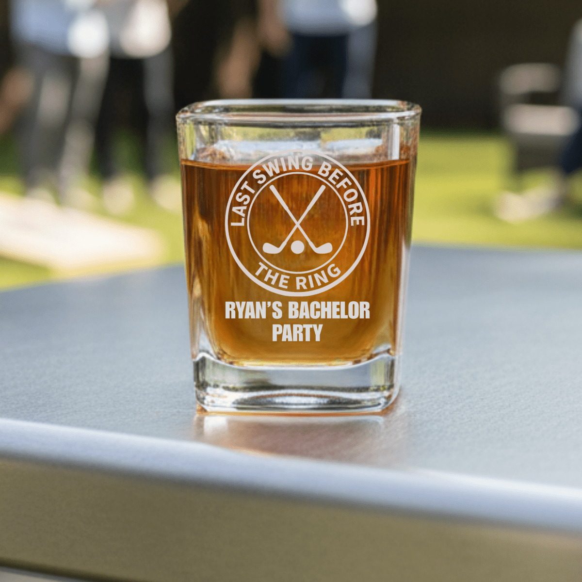 Glass of beer with 'Last Swing Before the Ring' and 'Ryan's Bachelor Party' text on a table outdoors.