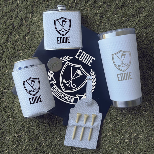 Groomsmen gift set with flask, tumbler, can cooler, and golf tees on grass.