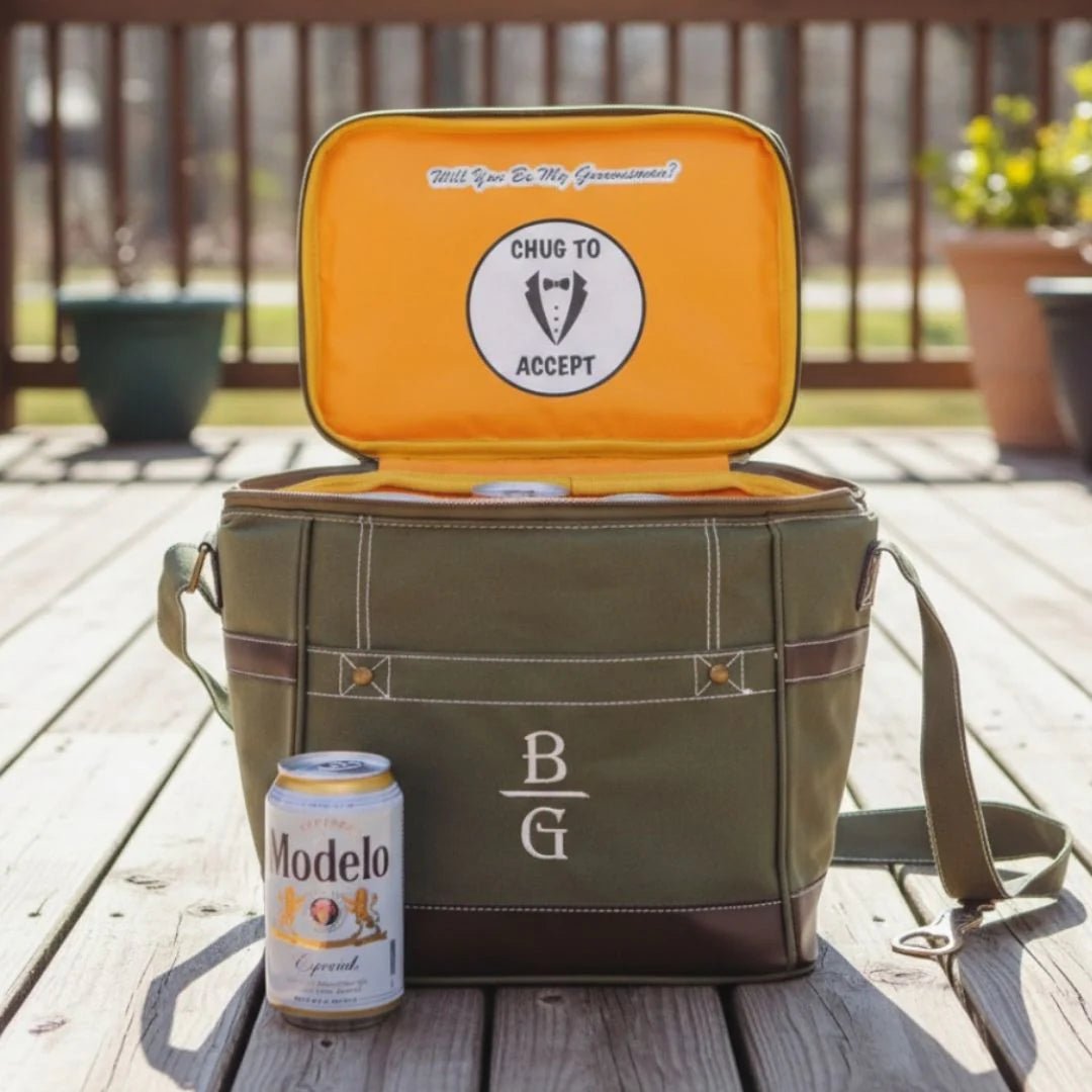 Green cooler bag with open lid showing 'Will You Be My Groomsman?' question, 'Chug to Accept' sticker, and a can of Modelo beer on a wooden deck.
