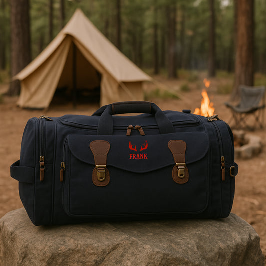 Black canvas duffle bag with brown leather accents, embroidered with sample name "FRANK" and red antler motif, displayed on rock with camping tent and campfire in background.