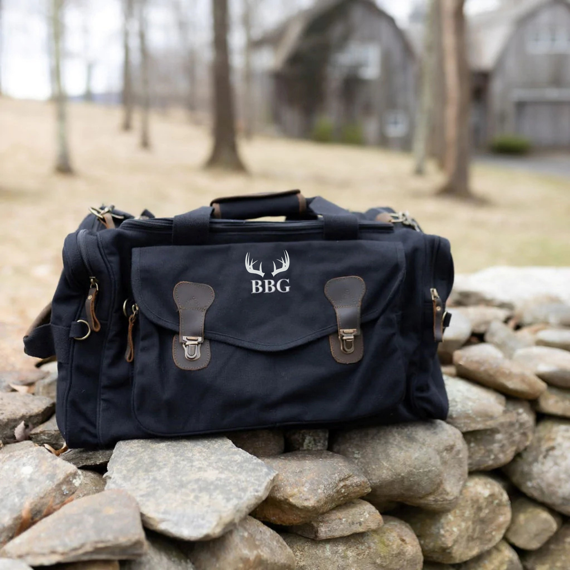 Navy canvas duffle bag with leather trim, embroidered with antler motif and sample initials "BBG" on front flap, displayed on stone wall outdoors