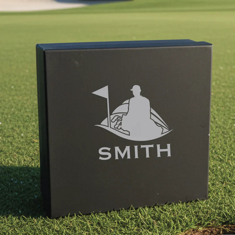 Black gift box with engraved golf flag and golfer silhouette, sample name "SMITH" engraved below, displayed on grass