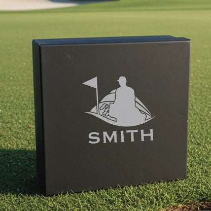 Black gift box with engraved golf flag and golfer silhouette, sample name "SMITH" engraved below, displayed on grass
