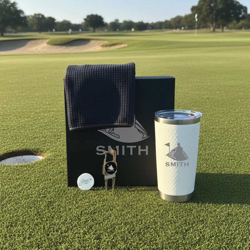 Golf-themed gift set with personalized tumbler engraved "SMITH", navy towel, divot tool, and golf ball on course green.