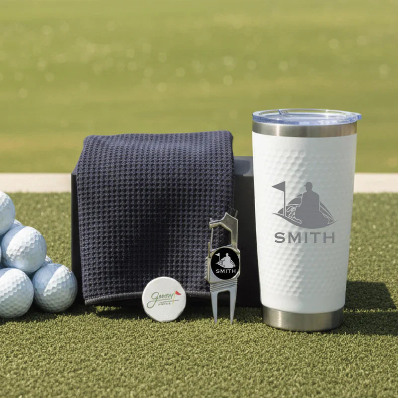 Golf gift set with navy towel, white insulated tumbler engraved with "SMITH" and golfer silhouette, divot tool, and golf balls on green turf background.