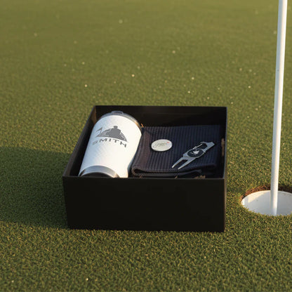 Black gift box containing white engraved tumbler with golf logo, dark pouch with divot tool, and small oval personalization sample on putting green.