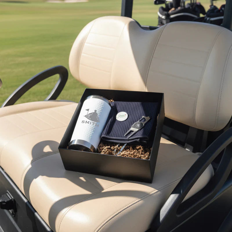 Black gift box containing engraved flask with "Smith" sample personalization, navy towel, and golf ball sitting on golf cart seat.