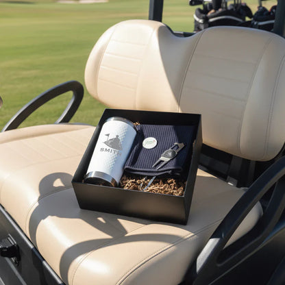 Black gift box containing engraved flask with "Smith" sample personalization, navy towel, and golf ball sitting on golf cart seat.