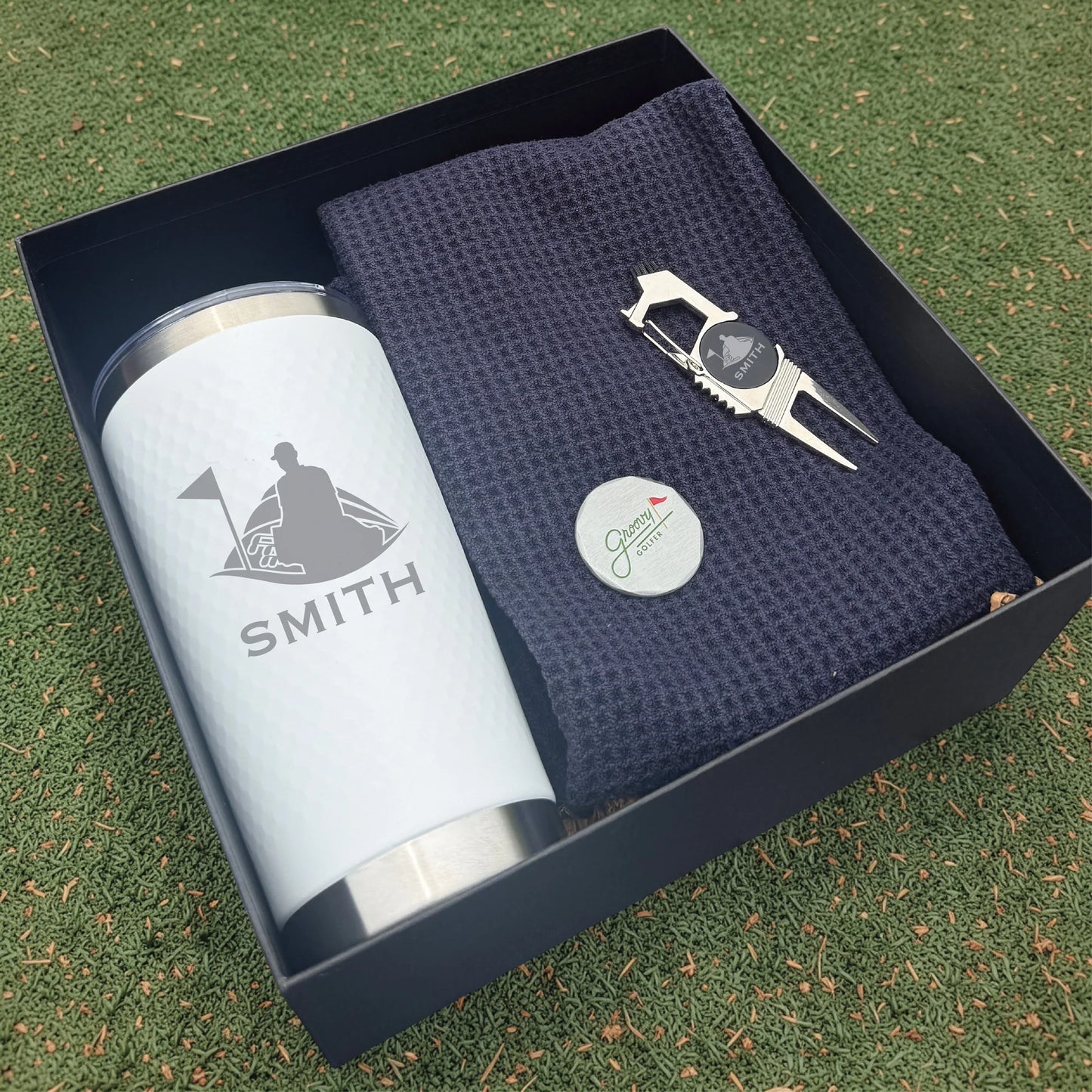 Golf-themed gift set with tumbler, towel, and divot tool in a box on grass.