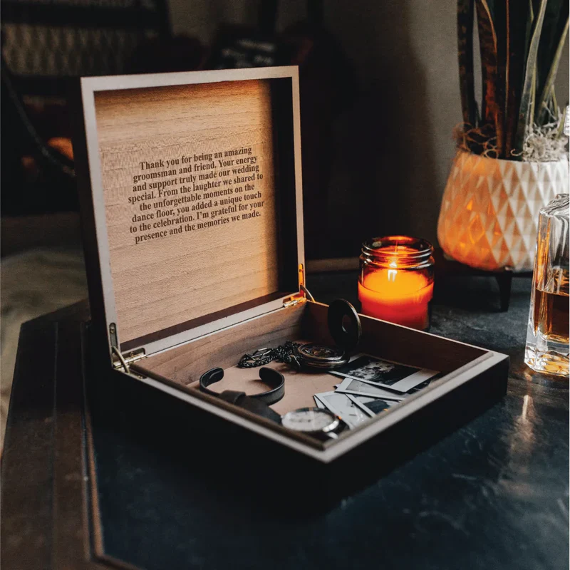 Open wooden keepsake box with engraved thank-you message sample inside, containing cufflinks and other small items on dark surface with candle and glasses nearby