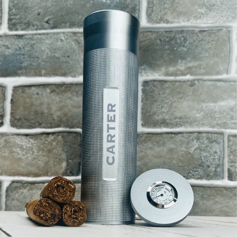 Engraved stainless steel flask with "CARTER" sample personalization, displayed with three cigars and a cigar humidor gauge against a brick wall backdrop.