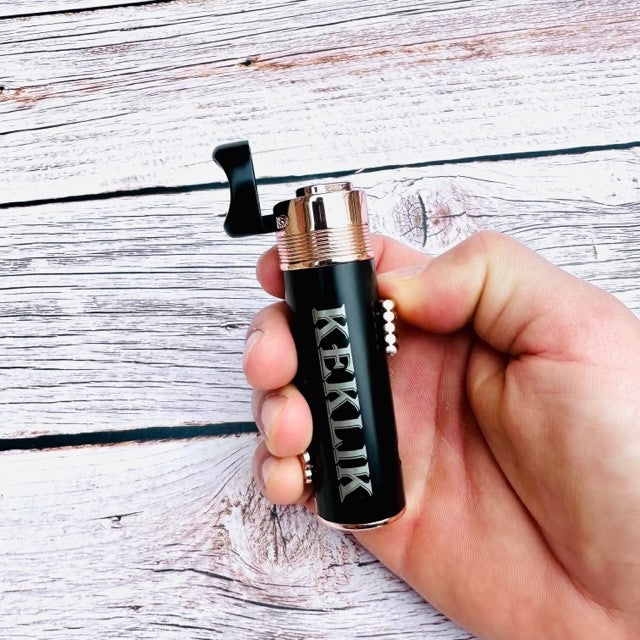 Hand holding black engraved cigar lighter with sample personalization, gold and silver trim, whitewashed wood background.