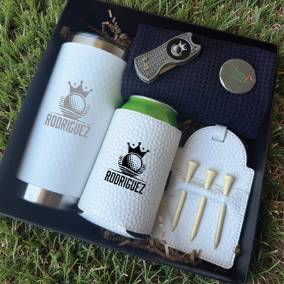 Black gift box containing white insulated tumbler and can cooler with sample "Rodriguez" engraving, golf divot tool, cigar cutter, and golf tee holder with wooden tees, displayed on grass.