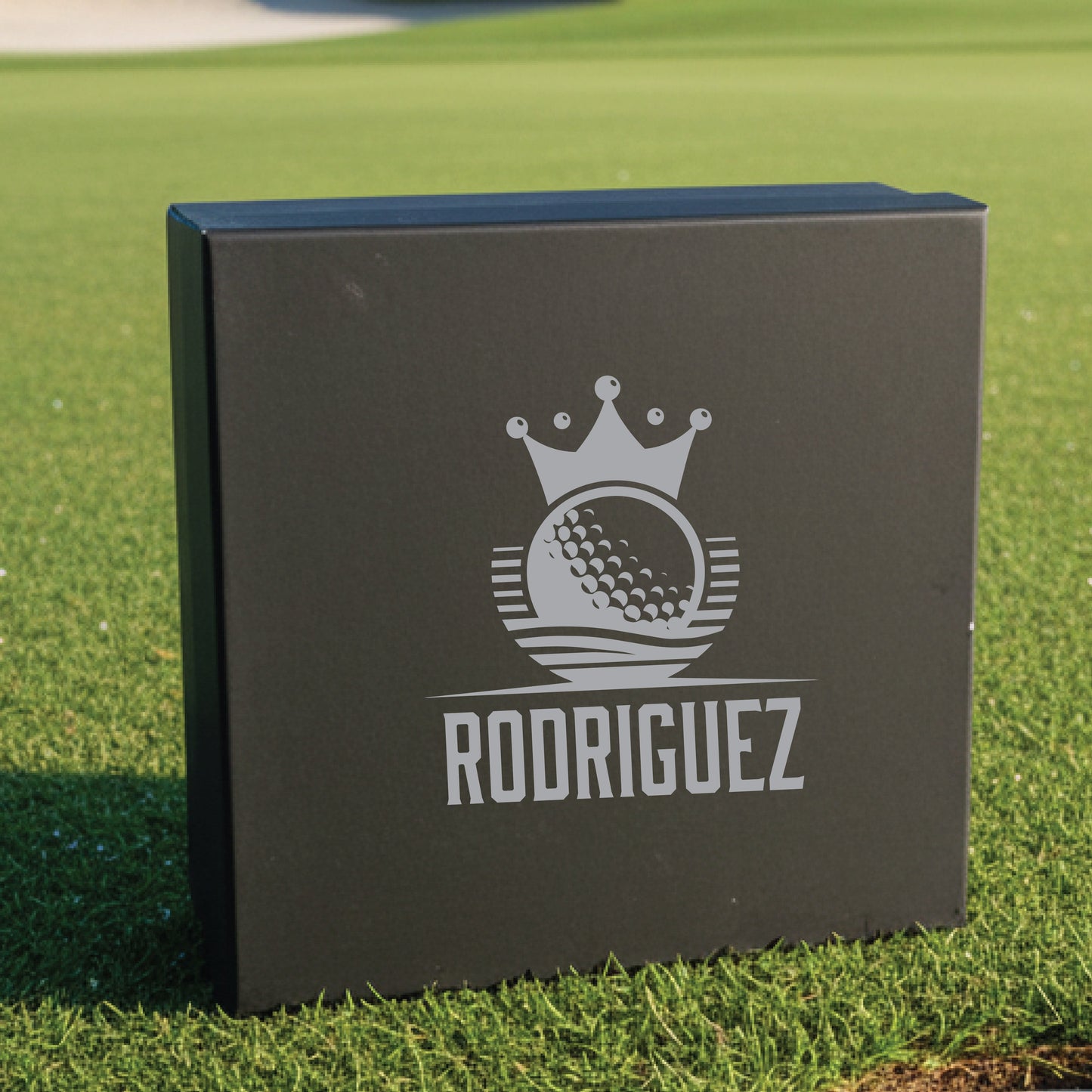 Black gift box with engraved golf crown logo and sample name "Rodriguez" displayed on grass background.