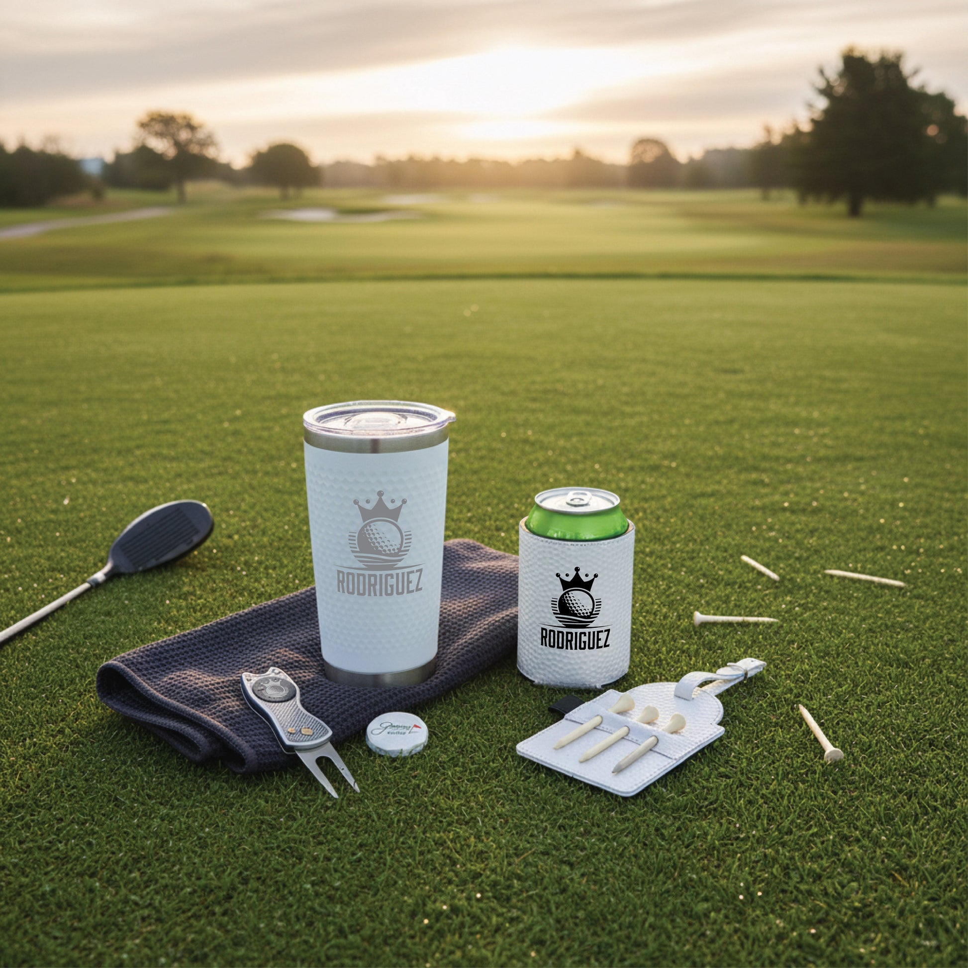 Golf accessories displayed on putting green with sample "Rodriguez" engraving on insulated tumbler and can cooler, including divot tool, ball marker, and tee set.