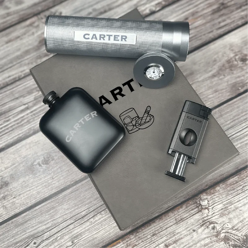 Engraved flask, cigar cutter, and lighter with "CARTER" sample personalization displayed on gray leather surface.