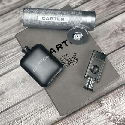 Engraved flask, cigar cutter, and lighter with "CARTER" sample personalization displayed on gray leather surface.