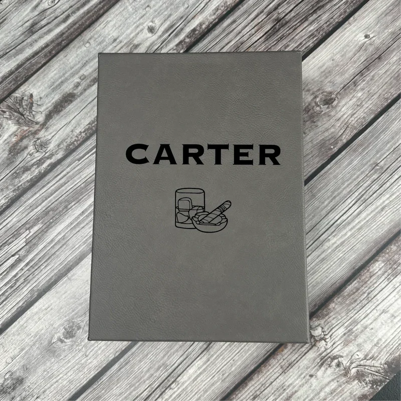 Gray leatherette box with "CARTER" engraved on cover and flask and cigar icons below, displayed on weathered wood surface