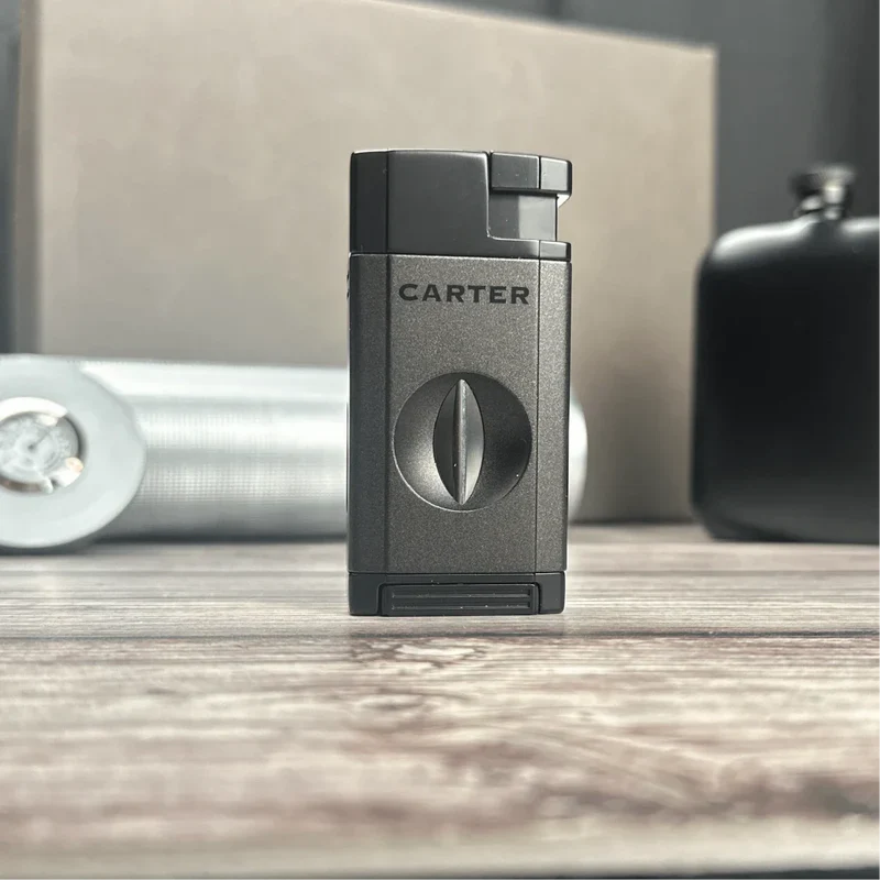Gunmetal gray Carter cigar lighter with engraved brand name and circular design detail, displayed on wooden surface with flask and cigar accessories in background
