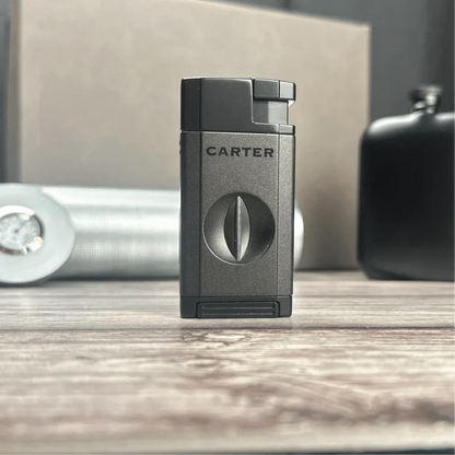 Gunmetal gray Carter cigar lighter with engraved brand name and circular design detail, displayed on wooden surface with flask and cigar accessories in background
