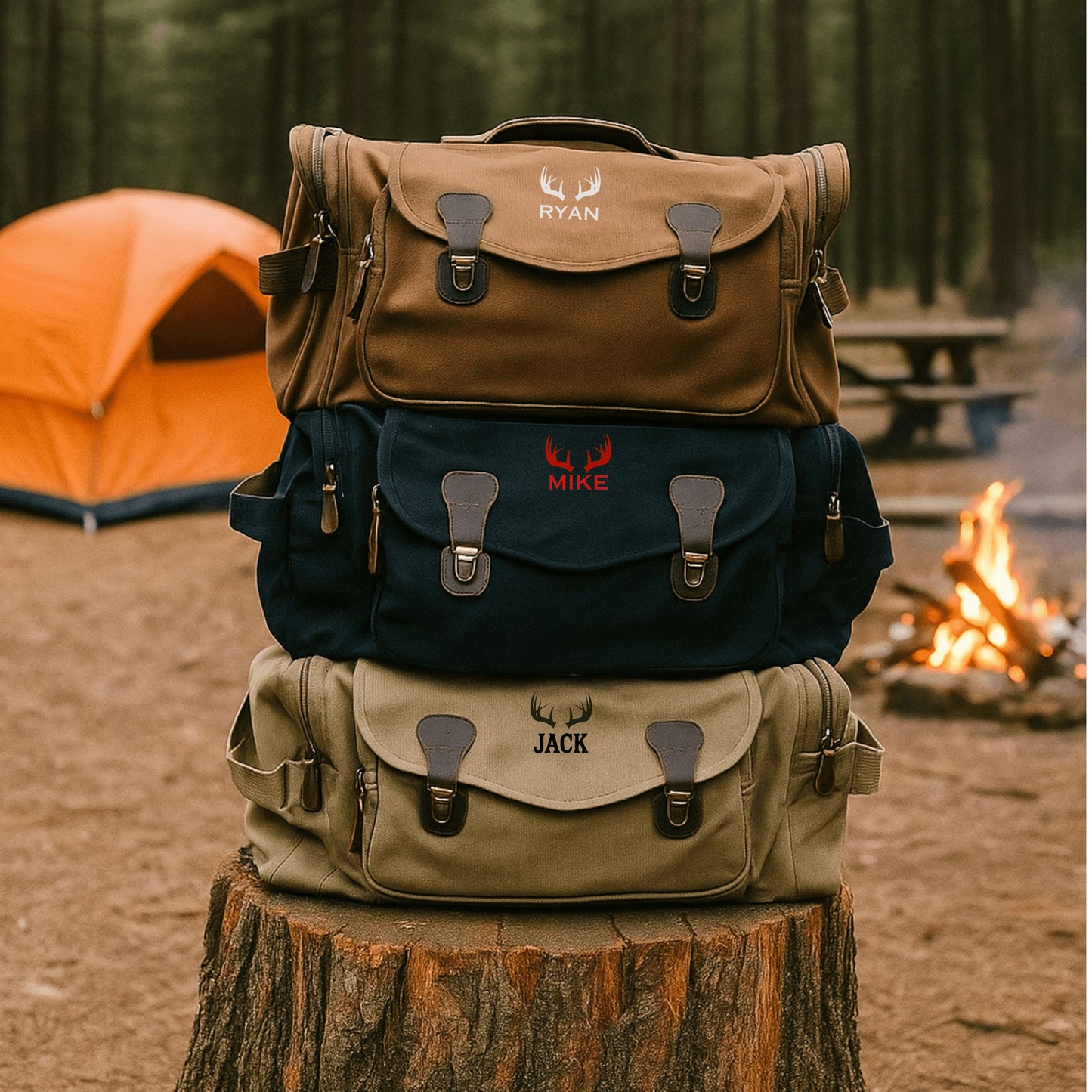 Three canvas duffle bags stacked on a wood log, each personalized with sample names "RYAN," "MIKE," and "JACK" embroidered with antler motifs in brown, navy, and khaki colors.