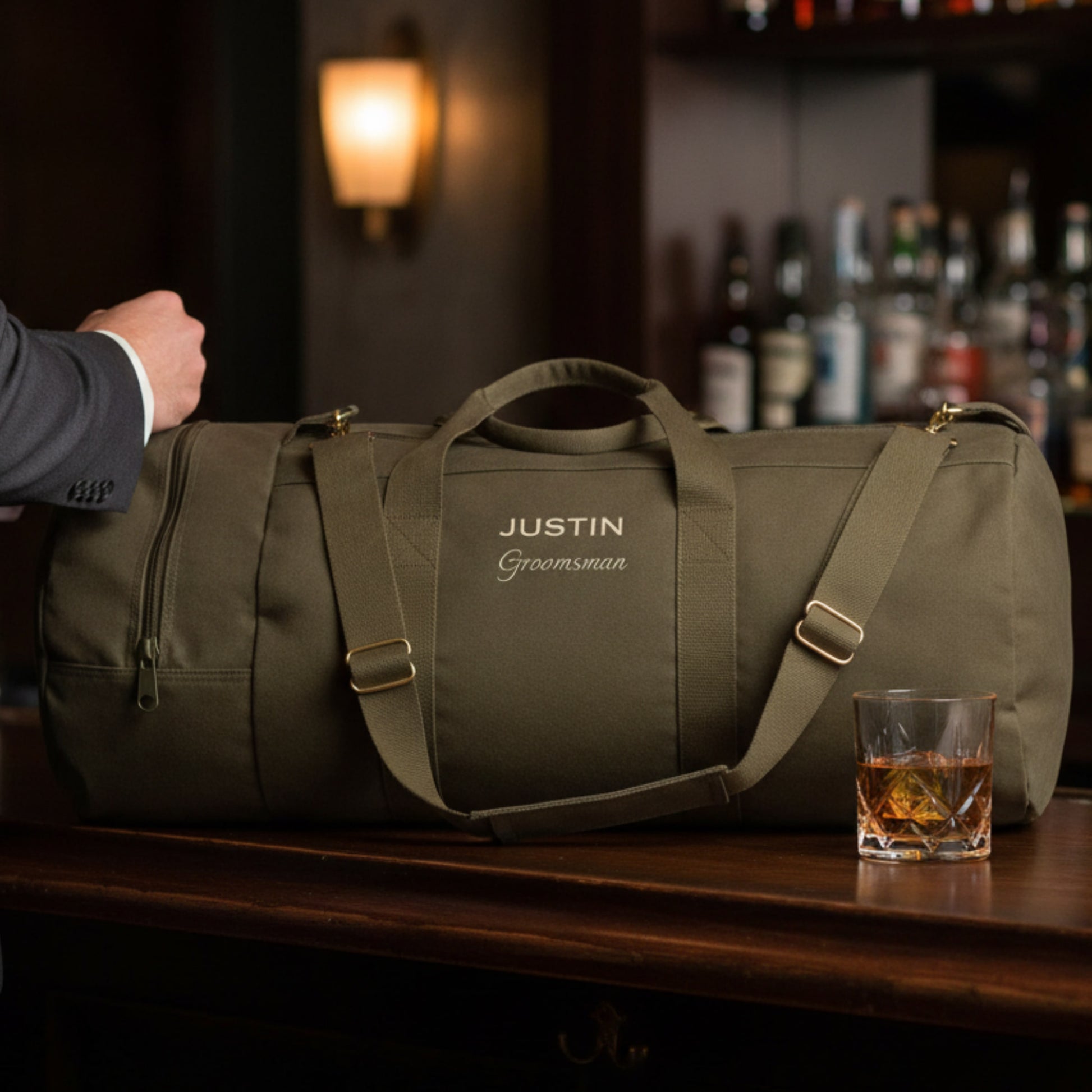 Olive canvas duffel bag with sample personalization reading "JUSTIN" and "Groomsman" displayed on a bar counter next to a whiskey glass