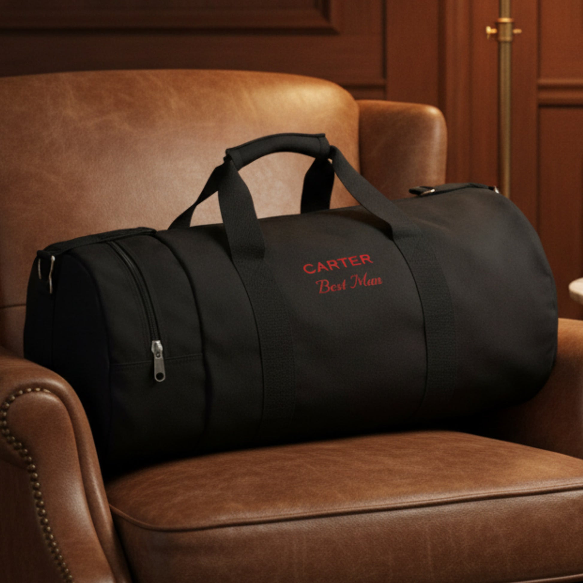 Black canvas duffle bag with sample personalization reading "CARTER" and "Best Man" in red embroidery, displayed on a tan leather chair