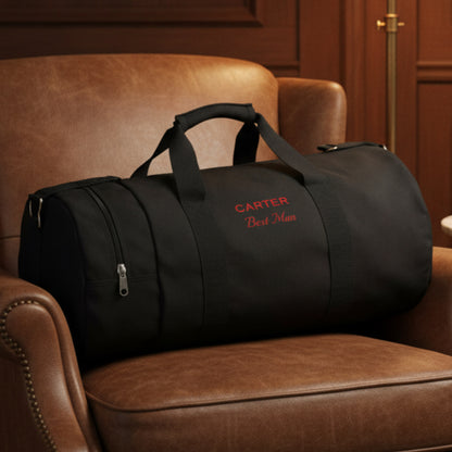Black canvas duffle bag with sample personalization reading "CARTER" and "Best Man" in red embroidery, displayed on a tan leather chair