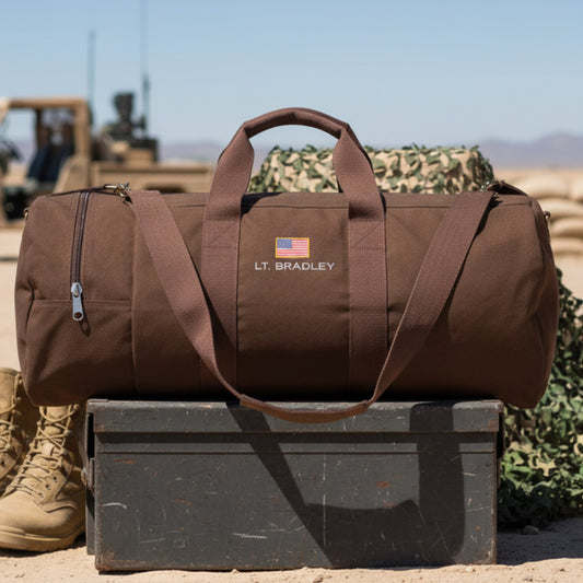 Brown canvas duffle bag with "LT. BRADLEY" and American flag embroidered on front, displayed on military equipment in desert setting.
