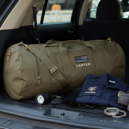 Tan canvas duffle bag with "CARTER" and blue line flag embroidered on side, displayed in car trunk with folded navy shirt, flashlight, and engraved rings.