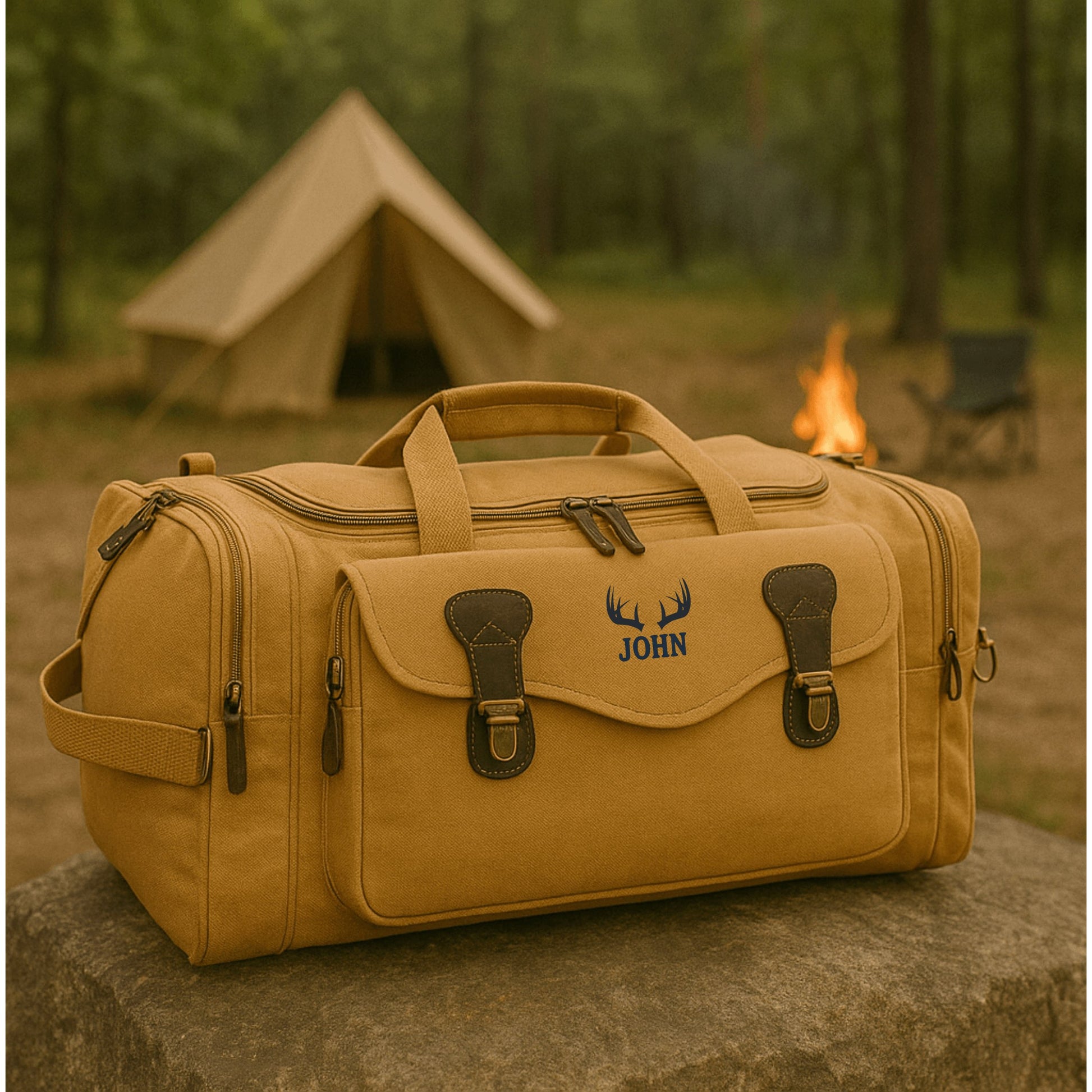 Khaki canvas duffle bag with personalized embroidered antler motif and sample name "JOHN" displayed in outdoor camping setting with tent and campfire.