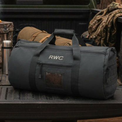 Black cylindrical duffle bag with RWC engraved on front, displayed in truck bed with outdoor gear and flask