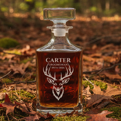 Engraved glass decanter with deer head design and sample personalization reading "CARTER GROOMSMAN 09/14/2026" filled with amber liquid, displayed outdoors on forest floor.