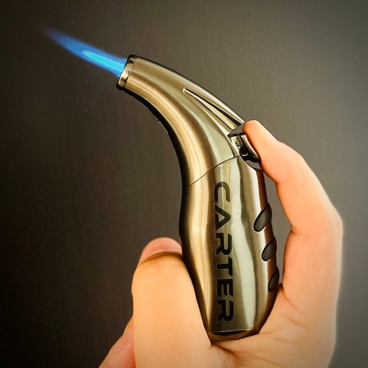 Gold-toned torch lighter with "Carter" engraved on side, held in hand with blue flame ignited, against neutral background.