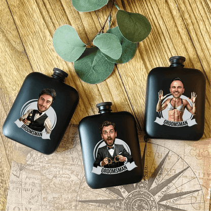 Three black engraved flasks with sample photo personalization showing different men and "Groomsman" text, arranged on wooden surface with eucalyptus leaves and vintage compass design.