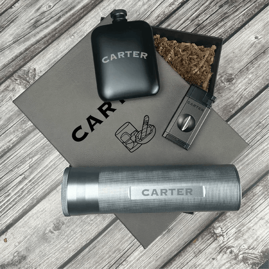 Black engraved flask, cigar cutter, lighter, and cork-lined pouch with sample "CARTER" personalization on gray leather mat.