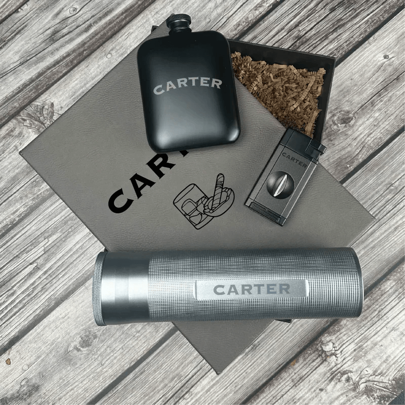 Black engraved flask, cigar cutter, lighter, and cork-lined pouch with sample "CARTER" personalization on gray leather mat.