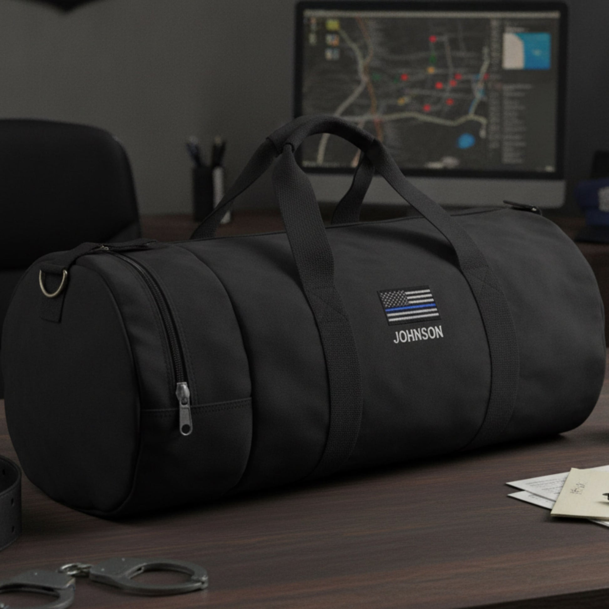 Black cylindrical duffle bag with sample personalization showing "JOHNSON" text and thin blue line flag emblem on front.