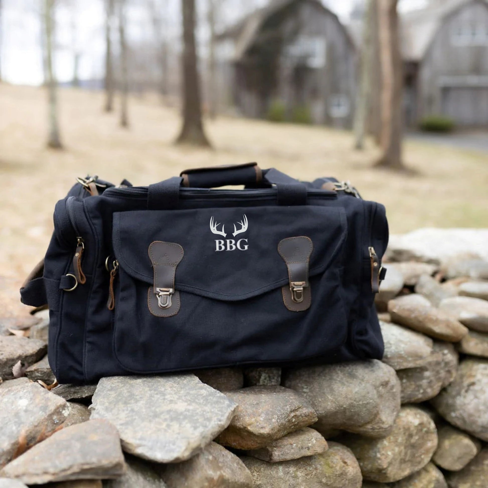 Navy canvas duffle bag with leather trim, embroidered with antler motif and sample initials "BBG" on front flap, displayed on stone wall outdoors