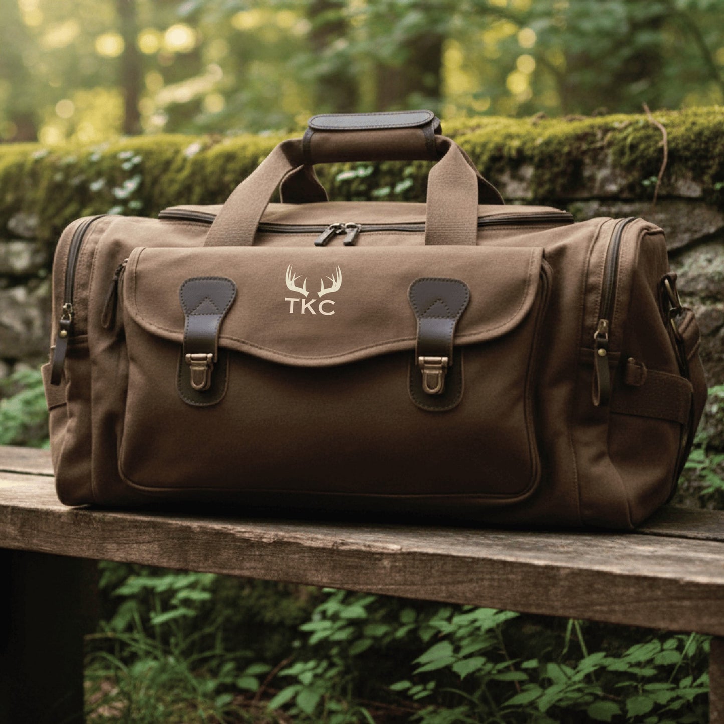 Brown canvas duffle bag with leather accents, embroidered with sample antler motif and "TKC" monogram on front flap, dual carry handles and side pockets visible