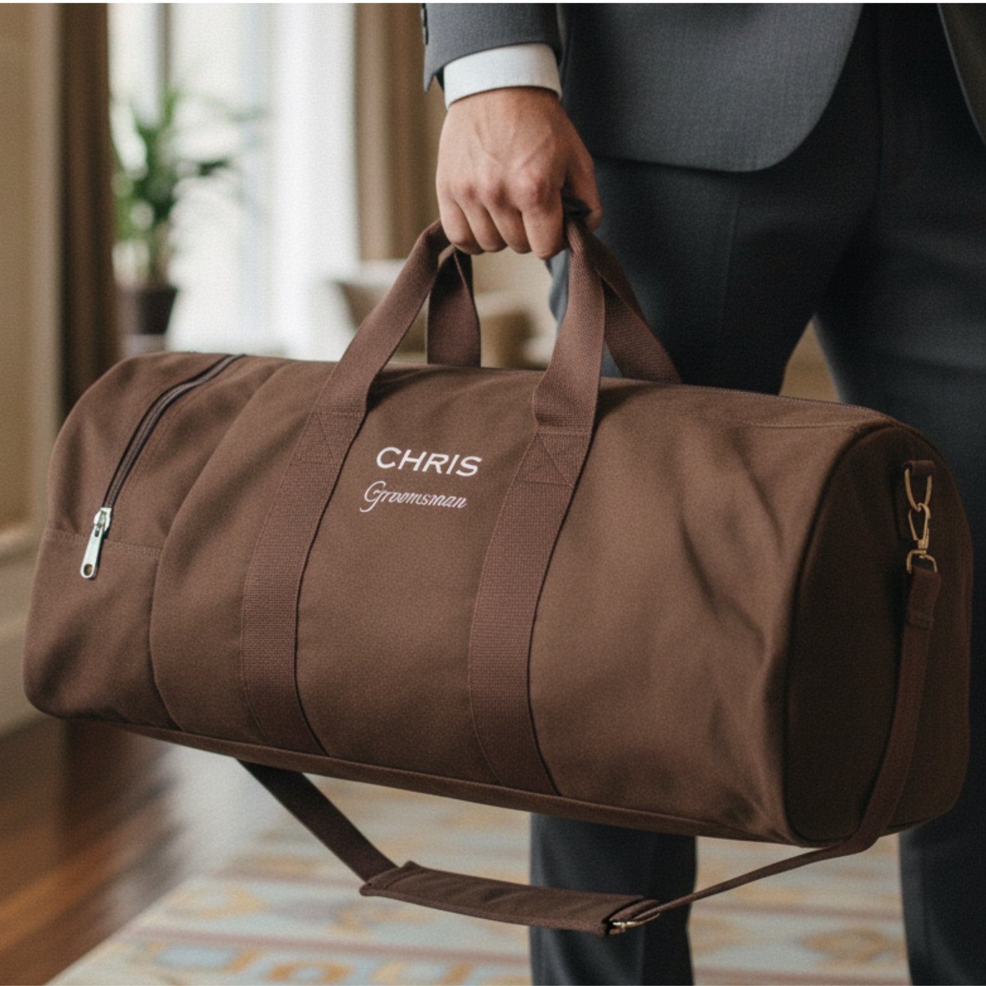 Brown canvas duffel bag with sample personalization reading "CHRIS" and "Groomsman" in white embroidery, held by man in gray suit.