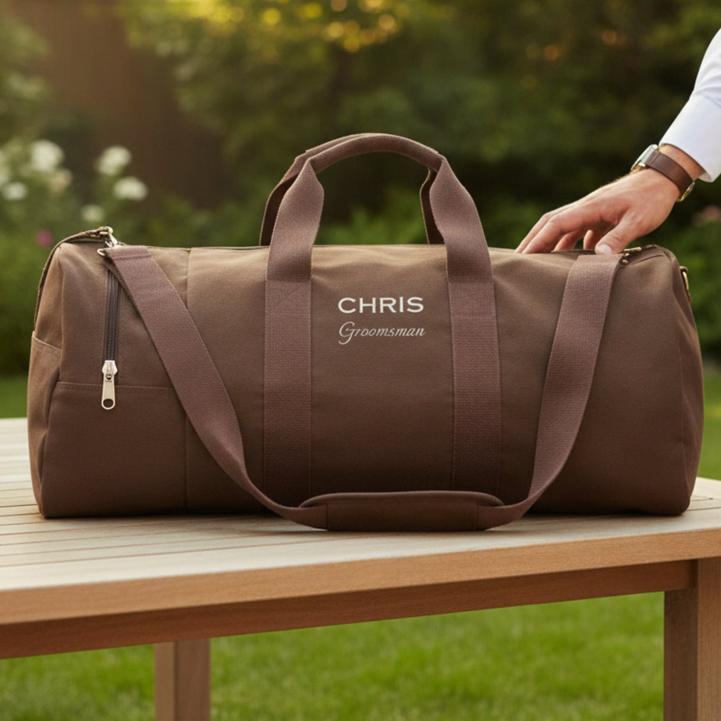 Taupe canvas duffel bag with sample personalization reading "CHRIS Groomsman" in white lettering, displayed on outdoor table with garden background