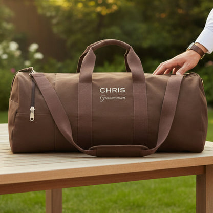 Taupe canvas duffel bag with sample personalization reading "CHRIS Groomsman" in white lettering, displayed on outdoor table with garden background