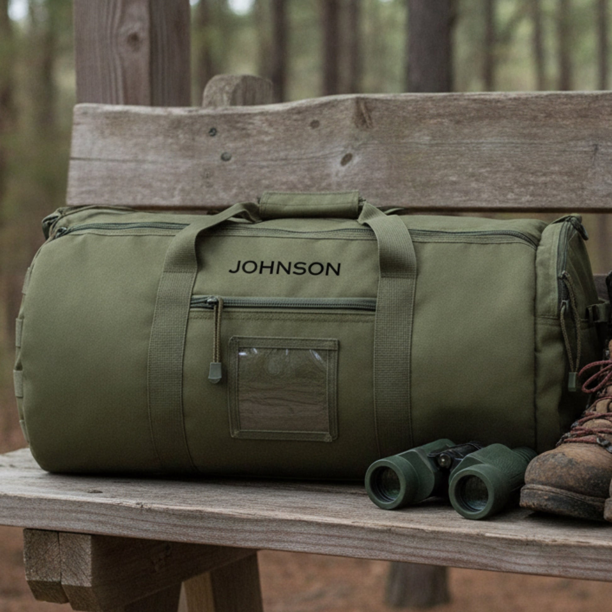 Olive green canvas duffle bag with "JOHNSON" engraved sample on front, displayed on wooden bench with binoculars and hiking boot nearby