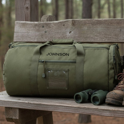 Olive green canvas duffle bag with "JOHNSON" engraved sample on front, displayed on wooden bench with binoculars and hiking boot nearby