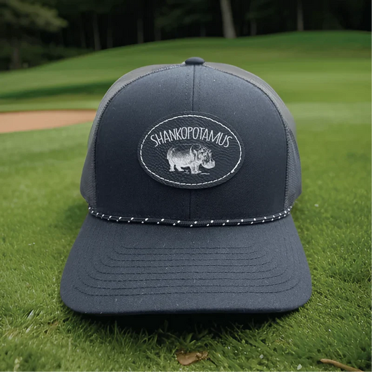 Dark gray trucker cap with embroidered oval patch reading "Shankopotamus" and hippo image, displayed on grass with baseball field background.